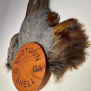 Può includere: Un design in legno di bossolo di fucile con le parole "SHOTGUN SHELL 12 GA" incise. Il design in legno è attaccato a una collezione di piume grigie e marroni.