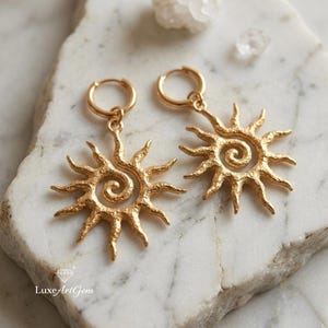 Puede incluir: Un par de pendientes de aro dorados. Cada pendiente presenta un diseño de sol con un centro en espiral. Los pendientes se exhiben sobre una superficie de mármol blanco y gris. Una joya.