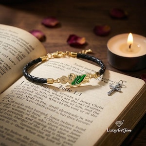 Può includere: Un braccialetto dorato con un cinturino in pelle nera, con un ciondolo a forma di santo e bandiera verde, è appoggiato su un libro aperto. Sono presenti anche un piccolo ciondolo a forma di croce d'argento e una candela accesa. Il libro mostra il testo "Giuda 1:1".