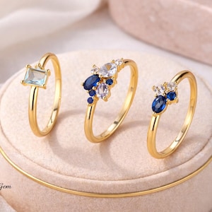 Puede incluir: Tres anillos de oro con engastes de piedras preciosas. Uno presenta una piedra azul claro rectangular, los otros grupos de piedras blancas y azules. Los anillos se exhiben sobre una superficie rosa suave.