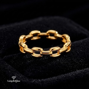 Puede incluir: Un anillo dorado con un diseño de eslabones de cadena. El anillo está hecho de un material metálico brillante y está sobre un fondo negro. El anillo es de talla 14.