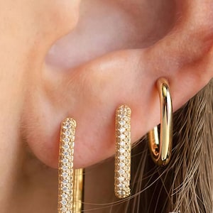 May include: Gold-toned hoop earrings in various styles. One pair features a rectangular shape encrusted with small, clear stones. Another pair is a simple, polished gold hoop. The earrings are worn on an ear, showcasing the jewelry's design.