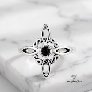 Puede incluir: Un anillo plateado con un diseño ornamental en forma de cruz. El anillo presenta una piedra negra central y redonda, y patrones intrincados y arremolinados. El anillo está sobre una superficie de mármol blanco.