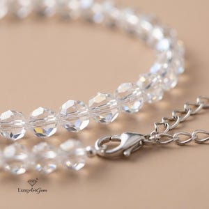 Puede incluir: Primer plano de una pulsera de cuentas de cristal transparente con un cierre y una cadena plateados. Las cuentas facetadas reflejan la luz, creando un efecto brillante. La pulsera está sobre un fondo neutro.