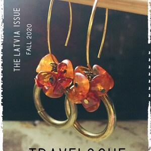 Op de afbeelding: Een paar gouden creolen met amberstenen. De oorbellen hangen aan een houten stok. De tekst "THE LATVIA ISSUE FALL 2020" is zichtbaar op de achtergrond.