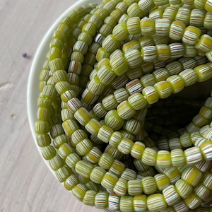 May include: A collection of small, round, translucent lime green glass beads arranged in a circular pile. Each bead features a series of thin, horizontal stripes in white, red, blue, and yellow, creating a detailed, multi-colored pattern across the surface.