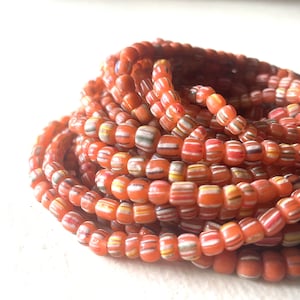 May include: A close-up of a pile of orange glass beads with white and multi-colored stripes. The beads are small and round, and they are arranged in a random pattern.