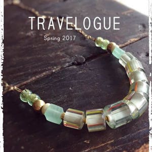 Könnte beinhalten: Eine grüne und goldene Halskette mit verschiedenen Perlen, darunter klare Glasperlen mit Streifen, grüne Glasperlen und goldene Perlen. Die Halskette liegt auf einer Holzoberfläche und der Text "TRAVELOGUE Frühling 2017" ist über der Halskette sichtbar.