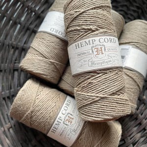 May include: A close-up shot of several spools of hemp cord. The cord is a natural tan color, tightly wound, and labeled with "HEMP CORD" and "Hemptique" branding. The spools are arranged in a woven basket.