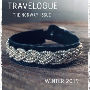 Op de afbeelding: Een zwarte leren armband met een zilveren kralenontwerp. De armband ligt op een houten oppervlak. De tekst "TRAVELOGUE THE NORWAY ISSUE WINTER 2019" is zichtbaar in de afbeelding.