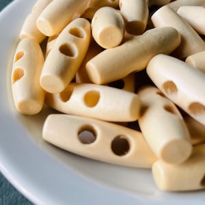May include: Close-up of a collection of light beige wooden toggle buttons. The buttons are oval-shaped with two or three holes and rounded ends. They are scattered on a white surface, suggesting a craft or sewing supply.