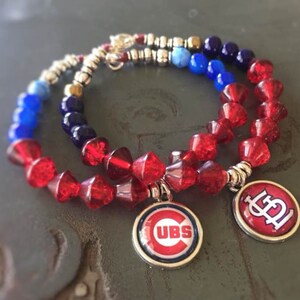 May include: Two beaded bracelets with red, blue, and silver beads. One bracelet has a charm with the Cubs logo, and the other has a charm with the Cardinals logo. The bracelets are likely for sports fans.