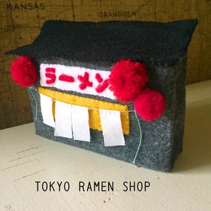 Puede incluir: Un modelo de tienda de ramen de Tokio hecho de fieltro con un techo negro, pompones rojos y un letrero amarillo que dice "Ramen" en caracteres japoneses. La tienda tiene cortinas blancas y un exterior gris.