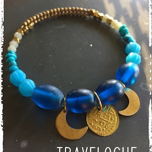 Op de afbeelding: Een armband met blauwe en gouden kralen. De armband heeft grote, ovale blauwe kralen, kleinere turkooizen kralen en goudkleurige kralen. Gouden halve maan- en muntbedels hangen aan de blauwe kralen. De tekst "TRAVELOGUE" en "FALL 2018" zijn zichtbaar.