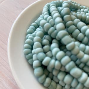 May include: A pile of light blue glass beads. The beads are small and round, and they have a slightly frosted finish.