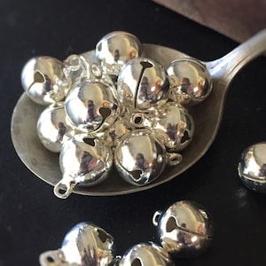 May include: A silver spoon filled with silver jingle bells. The bells are round with a heart-shaped opening.