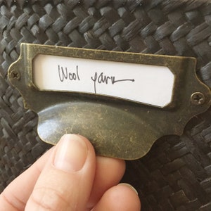 Könnte beinhalten: Gealterter Messing-Etikettenhalter mit dem handgeschriebenen Text "Wool yarn" auf einem weißen Etikett. Der Etikettenhalter ist an einem dunklen Webkorb befestigt. Der Etikettenhalter wird von einer Hand gehalten.