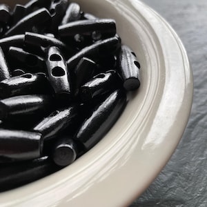 May include: A collection of black toggle buttons with a smooth, glossy finish. Each button is elongated and cylindrical in shape, featuring two centered holes for attachment. The buttons are gathered together in a light-colored bowl.