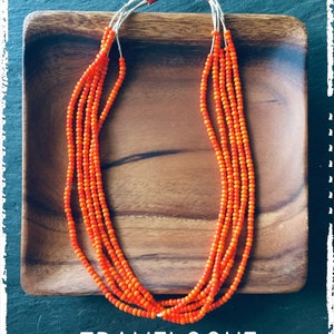 Op de afbeelding: Een meerstrengs ketting gemaakt van oranje kralen op een houten bord. De ketting is gemaakt van meerdere strengen kleine, ronde kralen. De kralen zijn fel oranje van kleur. De ketting ligt op een houten bord. De tekst "TRAVELOGUE SUMMER 2020" is onder de ketting gedrukt.