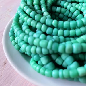 May include: A pile of turquoise-colored beads, tightly coiled on a white ceramic plate. The beads are small, rounded, and have a matte finish. The plate is slightly off-white, and the background is a light pink.