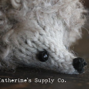 May include: A close-up of a gray knitted hedgehog with black eyes and a black nose. The hedgehog is lying on a wooden surface.