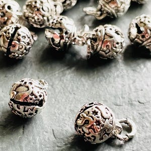 Könnte beinhalten: Nahaufnahme mehrerer silberfarbener Filigranperlen. Die Kugeln haben aufwendige, durchbrochene Muster. Einige Perlen sind durch kleine Ketten verbunden. Die Perlen sind auf einer grauen Oberfläche verstreut.