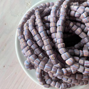 May include: A collection of light purple beads with colorful striped patterns, arranged in a circular shape on a white plate. The beads have a cylindrical shape and are tightly strung together, creating a textured and visually appealing design.