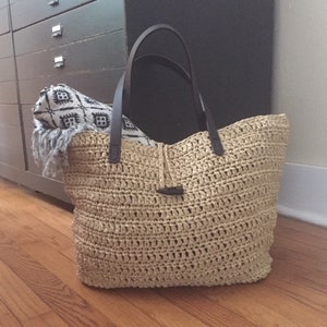 May include: A large, beige straw tote bag with brown leather handles. The bag is made of a woven material and has a small, brown leather buckle on the front.
