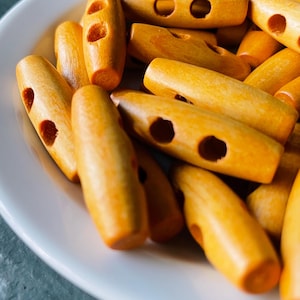 May include: A collection of light orange wooden toggle buttons with two holes, displayed on a white plate. The buttons have a smooth, rounded shape and are made of wood. The buttons are a warm, natural color.