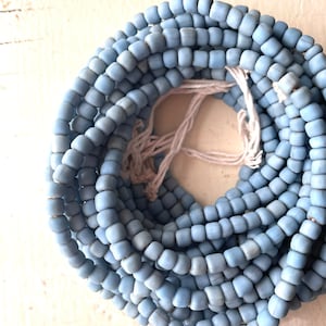 May include: A collection of light blue, square beads strung on white string, forming a coiled bundle. The beads are closely arranged, creating a dense pattern. The beads have a matte finish and a slightly aged look, possibly vintage or antique.