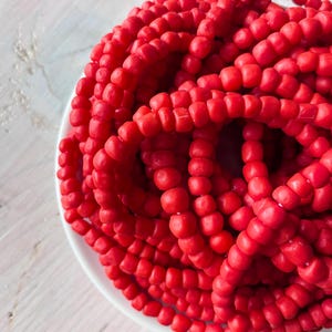 May include: A pile of bright red, round beads, possibly glass or plastic, are arranged in a white bowl. The beads are strung together, forming long strands. The background is a light-colored, textured surface.