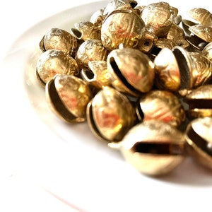 Puede incluir: Un montón de pequeñas campanas doradas con una superficie texturizada. Las campanas están dispuestas en un patrón aleatorio sobre una superficie blanca.