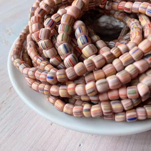 May include: Close-up of a collection of pink and multicolored striped beads. The beads are cylindrical and arranged in a circular pattern, resting in a white dish. The stripes include shades of red, blue, yellow, and green.