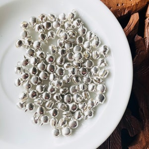 May include: A collection of small, silver-colored bells scattered on a white, round plate. The bells have a rounded shape with a small loop at the top. The plate is set against a dark wooden background, creating a contrast of textures and colors.