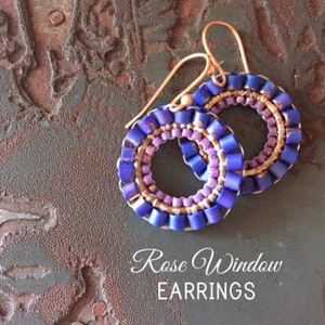 Könnte beinhalten: Ein Paar violette und goldene Perlenohrringe mit einem Rosettenmuster. Die Ohrringe liegen auf einer braunen, strukturierten Oberfläche. Rose Window EARRINGS.