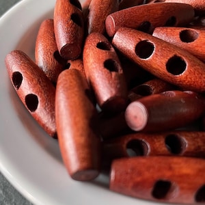 May include: Close-up of a collection of reddish-brown wooden toggle buttons. Each button is oval-shaped with two holes and a smooth, polished finish. The buttons are in a white bowl, suggesting they are ready for use in sewing or crafting projects.