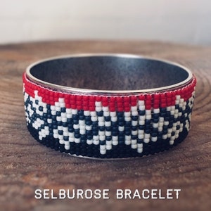 May include: A beaded bracelet with a black, white, and red geometric pattern. The bracelet is made of silver metal and has a simple, elegant design.