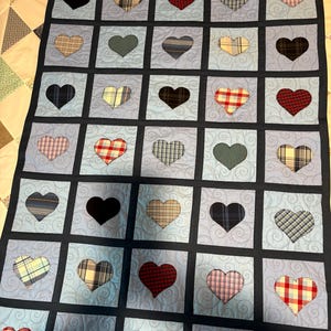 May include: A quilt featuring a grid of light blue squares with a swirling pattern, each containing a heart-shaped fabric appliqué. The hearts display various plaid, checkered, and solid patterns in colors including red, blue, black, and tan, all framed by dark navy blue sashing.