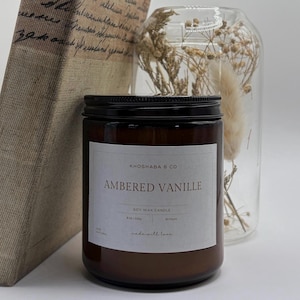 May include: A brown glass jar candle with a black lid and a white label that reads "AMBERED VANILLE" from Khoshaba & Co. The candle is 8 oz / 226g and is next to a clear glass jar with dried flowers and a book.