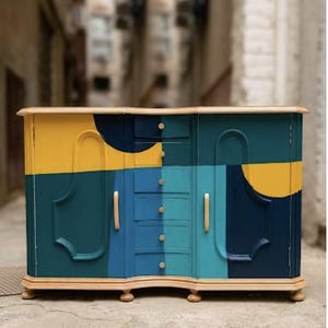 May include: A wooden sideboard featuring a painted geometric design in shades of yellow, teal, and dark blue. The cabinet includes two side doors with light-colored vertical handles and a central column of six drawers with small round knobs.