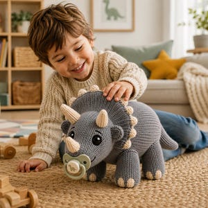 May include: A grey knitted triceratops stuffed animal featuring a large frill with cream-colored edges, three cream-colored horns, and a light green pacifier in its mouth. The pacifier has a small dinosaur graphic printed on the center button.