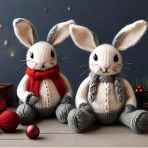 May include: Two knitted bunny figurines with white bodies, grey and white ears, and grey legs. One bunny wears a red scarf, the other a grey scarf. The bunnies are sitting on a surface with Christmas ornaments and a lit candle.