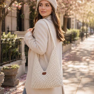 May include: A cream-coloured crocheted hobo bag with a textured diamond pattern and a single braided shoulder strap. The bag features a small, rectangular brown leather-like patch centred on the front exterior, showcasing a simple, functional design.