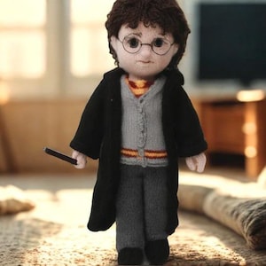 May include: A handmade doll of a character with brown hair, round glasses, and a black robe. The doll wears a gray sweater with a red and gold striped tie and holds a black wand. The doll is standing on a patterned surface.