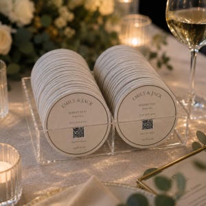 May include: Two stacks of round, off-white coasters held in a clear acrylic organiser. Each coaster features the text "EMILY & JACK", "MARRY YOU", "Bruno Mars", "PLAY OUR SONG", a QR code, "Our First Dance", and the date "14.08.2026" printed in dark grey.