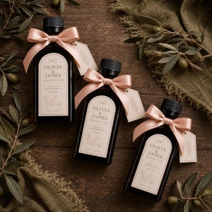 May include: Three small, dark glass bottles with black caps, each tied with a blush pink ribbon and a tag. The labels read "Olivia & James Engagement Favor" with a floral design and date. Olive branches and olives are scattered around.