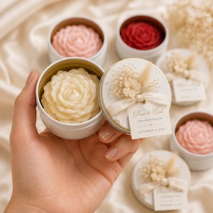 Puede incluir: Una lata redonda de metal blanco que contiene una vela con forma de flor de color crema. La tapa está decorada con flores secas, una cinta color crema y una etiqueta que dice Olivia & Noah, ENGAGEMENT FAVOR, SEPTEMBER 14, 2026. Hay otras latas.