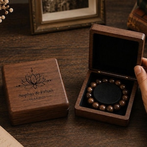 May include: Two square dark wood boxes with hinged lids. One box is closed, featuring a laser-engraved lotus flower design with the text "Sophia & Ethan" and "06-18-2026". The open box contains a circular dark brown beaded bracelet on a black insert.