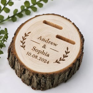 May include: A rustic wooden ring holder, cut from a tree slice, with two slots for rings. The surface is engraved with the names "Andrew & Sophia" and the date "10.08.2024", framed by a laurel wreath design. The bark edge is visible.