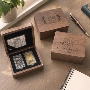 Puede incluir: Tres cajas de madera oscura con tapas abatibles. Una caja está abierta, mostrando una barra de plata con la inscripción one ounce 999 fine silver y una barra de oro sobre un inserto de terciopelo negro. Las cajas cerradas tienen grabados J|M EST. 2024 y BUILT TO LAST EST. 2024.